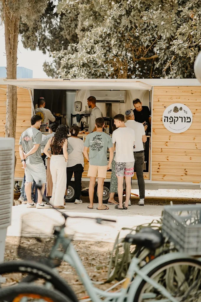 עגלת הקפה של מורקפה בכניסה לקיבוץ עלומים, אורחים בתור מתחת לעצים